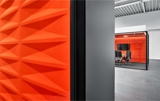 Detailaufnahme einer verglasten Besprechungsbox in der Produktionshalle von ›Grohe Technology‹ mit markanter, strukturiert ausgeführter Wandoberfläche in Orange als Teil des New-Work-Konzepts innerhalb der industriellen Arbeitsumgebung, geplant und realisiert von arc+ Architekten.